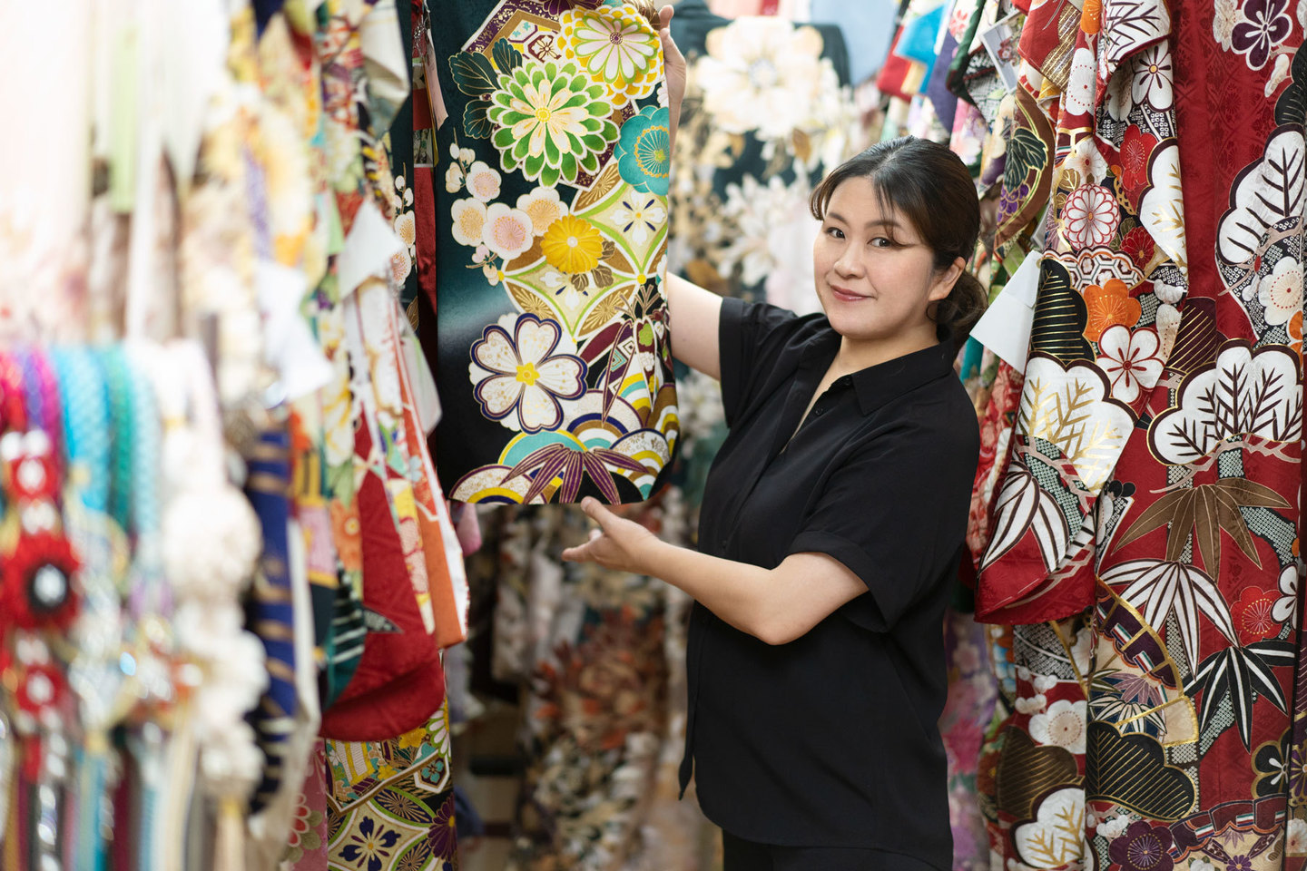 三原市の冨士写真館で振袖選びをサポートする専門スタッフ。数多くの振袖に囲まれた店内で、お嬢様にぴったりの一着を提案する、親しみやすくプロフェッショナルな接客風景。