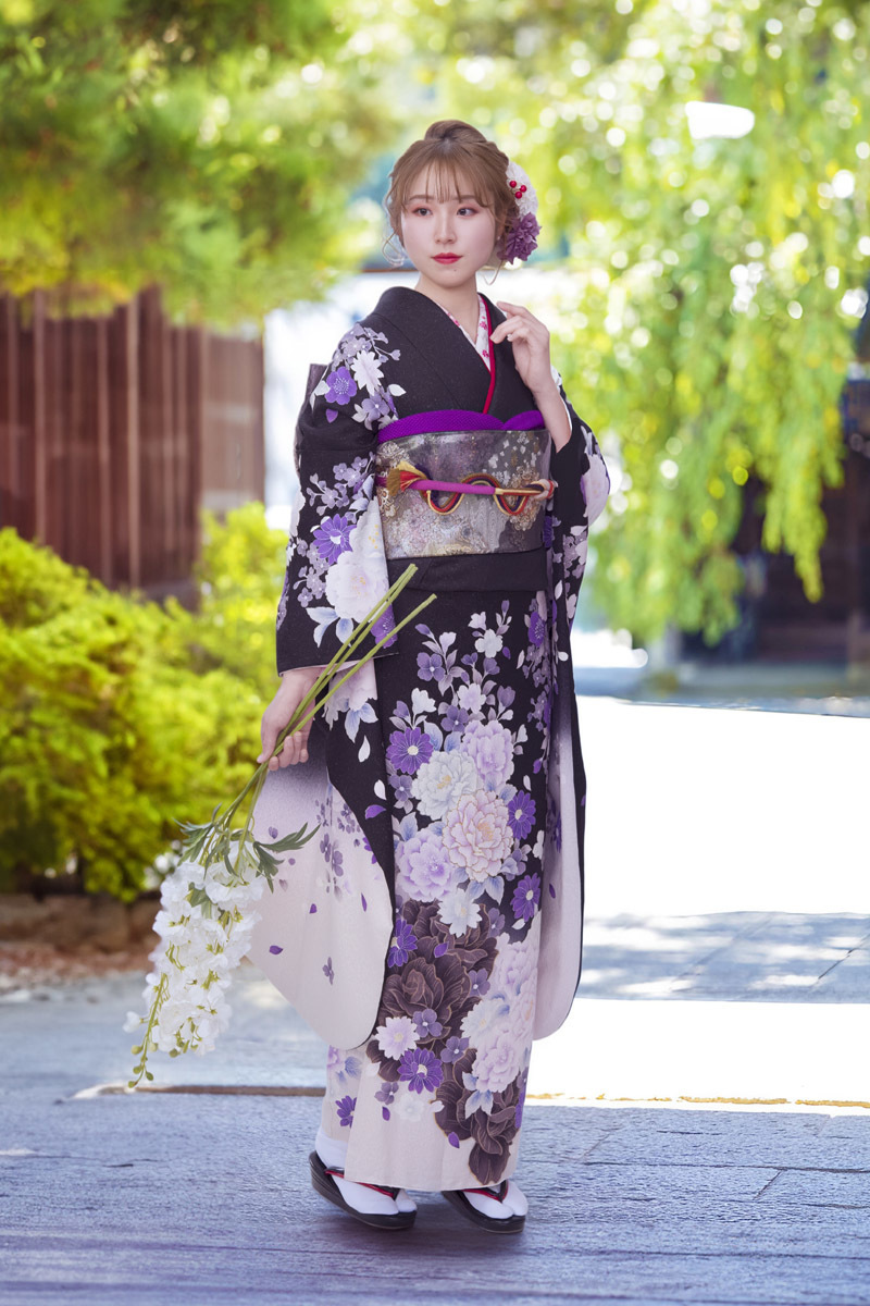 黒から白へのグラデーションが美しいモダンな振袖。紫や淡いピンクの牡丹が咲き誇り、屋外の自然光に映える。凛とした美しさとエレガントさを兼ね備えた、二十歳の門出を彩るスタイル。